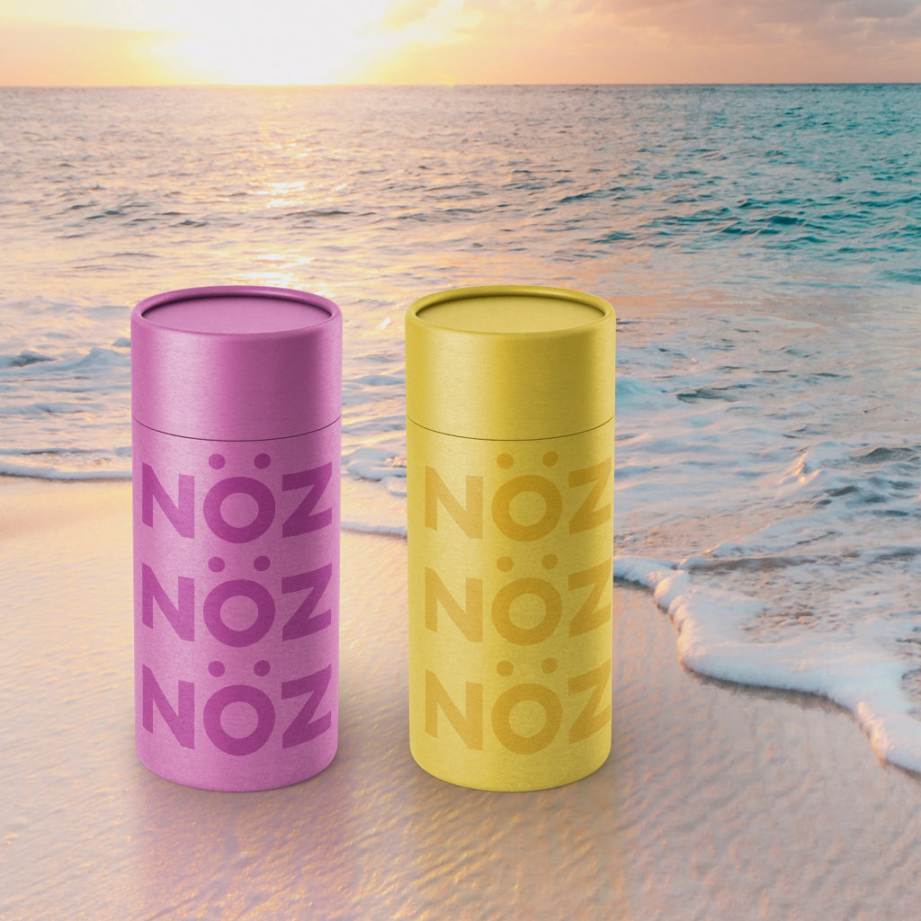 Nöz’s reef safe and vegan sunscreen sticks in the colors neon yellow and purple, on a beach background.